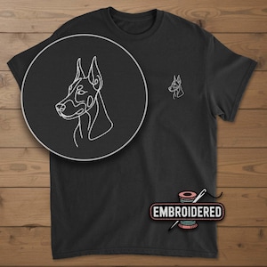 May include: Black t-shirt with a white embroidered outline of a Doberman Pinscher dog on the chest and a larger version in a circular inset. The word "EMBROIDERED" is printed on a pink spool of thread.