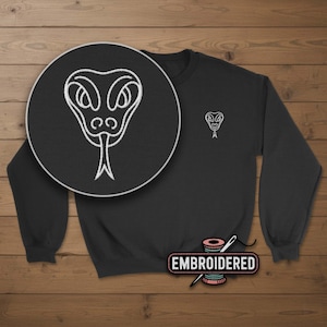 May include: Black sweatshirt with a white embroidered snake design on the chest and a larger, detailed snake head in a circular inset. The word "EMBROIDERED" is displayed on a label with a needle and thread.