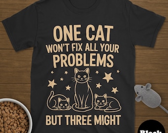 May include: Black t-shirt with a cat-themed graphic. The design features three cats and the text "ONE CAT WON'T FIX ALL YOUR PROBLEMS BUT THREE MIGHT". A bowl of cat food and a toy mouse are also visible.