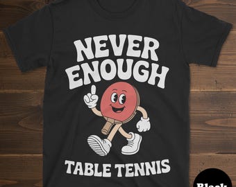 Puede incluir: Camiseta negra con el texto blanco "NEVER ENOUGH TABLE TENNIS". Una raqueta de tenis de mesa de dibujos animados con una cara sonriente y brazos que apuntan hacia arriba. La camiseta es de material suave.