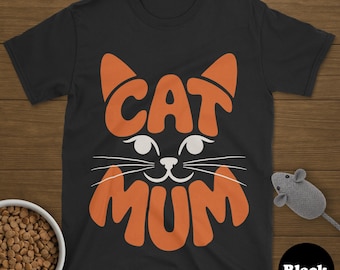 Può includere: T-shirt nera con un design a forma di faccia di gatto arancione. La parola "CAT" forma le orecchie del gatto e "MUM" forma la bocca. Sono visibili anche una ciotola di cibo per gatti e un topo giocattolo.