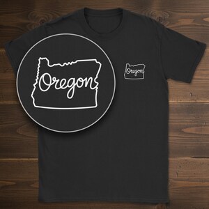 Puede incluir: Una camiseta negra de manga corta con un pequeño contorno blanco del estado de Oregón en el pecho. Dentro del contorno del estado, la palabra Oregon está escrita en letra cursiva blanca. Un recuadro circular muestra una vista ampliada.
