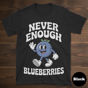 May include: Black t-shirt with the text "NEVER ENOUGH BLUEBERRIES" in white. A cartoon blueberry character with a smiling face and waving hand is featured in the center. The shirt is a crew neck.