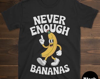 May include: Black t-shirt with the text "NEVER ENOUGH BANANAS" in white, above a cartoon banana character. The banana is yellow with white gloves and shoes, and is pointing upwards. A black circle with the word "Black" is in the bottom right corner.