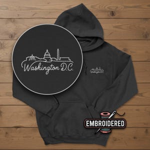 May include: Dark gray hoodie with a white embroidered design of the Washington D.C. skyline. The hoodie has a front pocket, a hood, and the words "Washington D.C." embroidered on the chest. The word "EMBROIDERED" is also visible.