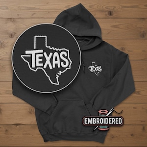 May include: A dark gray hoodie with a white embroidered outline of the state of Texas and the word "TEXAS". The hoodie has a front pocket and a hood. A close-up shows the embroidery detail. The word "EMBROIDERED" is on a label.
