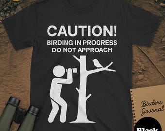 May include: Black t-shirt with white text and graphic. The text reads "CAUTION! BIRDING IN PROGRESS DO NOT APPROACH." A stick figure is looking through binoculars at a bird perched on a tree branch. A notebook and binoculars are in the image.