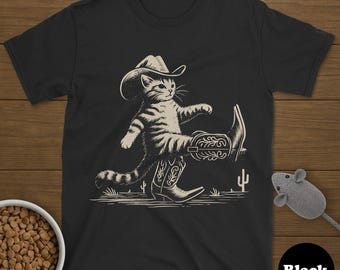 May include: Black t-shirt featuring a cartoon cat wearing a cowboy hat and boots, walking with a confident stride. The cat illustration is in a beige color, set against a black background. A cactus and a mouse toy are also visible.