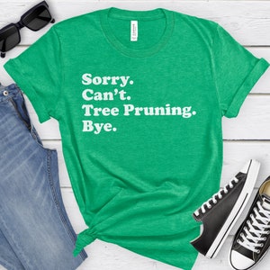 May include: A green t-shirt with the text "Sorry. Can't. Tree Pruning. Bye." in white. The shirt is styled with blue jeans, black sunglasses, and black and white sneakers on a white wooden surface.