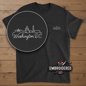 May include: Black t-shirt with white embroidered skyline of Washington D.C. and the text "Washington D.C." in a circular design. A smaller version of the design is on the upper right chest. The word "EMBROIDERED" is below the shirt.