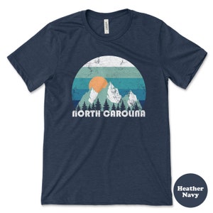 May include: A heather navy blue t-shirt with a vintage-style graphic of mountains, a sunset, and the text "NORTH CAROLINA".