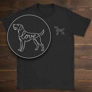 May include: A black short-sleeved t-shirt featuring a white line art graphic of a dog standing in profile. The word love is integrated into the dog's body outline. A larger circular inset shows the same graphic in greater detail.