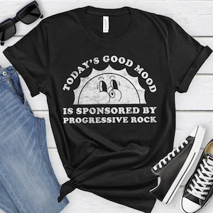 May include: Black t-shirt with the text "TODAY'S GOOD MOOD IS SPONSORED BY PROGRESSIVE ROCK" in white. The graphic features a smiling sun. The shirt is paired with blue jeans, sunglasses, and black sneakers.