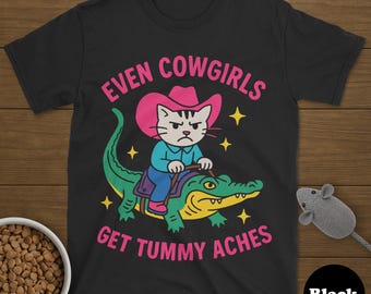 Peut inclure: T-shirt noir avec un dessin d'un chat portant un chapeau de cow-boy rose sur un alligator. Le texte "EVEN COWGIRLS GET TUMMY ACHES" est en rose. Des étoiles jaunes sont dispersées autour de l'image. Un cercle noir avec le mot "Black" est en bas à droite.