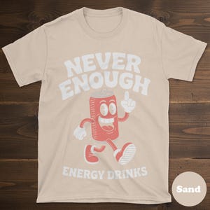 May include: Sand-colored t-shirt with the text "NEVER ENOUGH ENERGY DRINKS" in white. The graphic features a cartoon energy drink can with a smiling face, arms, and legs, pointing upwards. The can is red with white accents.