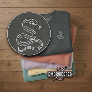 May include: A stack of folded sweatshirts in various colors, including gray, peach, lavender, yellow, and teal. The top sweatshirt is gray and features a small embroidered snake design. A larger circular image shows a detailed snake embroidery. The word "EMBROIDERED" is also visible.