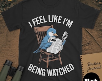 May include: Black t-shirt with the text "I FEEL LIKE I'M BEING WATCHED". The shirt features a blue bird reading a newspaper while sitting in a wooden chair. A notebook with the text "Birders Journal" is on the right.