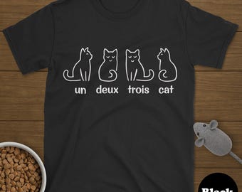 May include: Black t-shirt featuring a white graphic of four cat illustrations and the words "un deux trois cat". A bowl of cat food and a toy mouse are in the image. The t-shirt is black.
