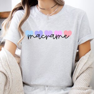 May include: A light gray t-shirt featuring the word "macrame" in a black script font. Above the word are five pastel-colored hearts in blue, purple, lavender, pink, and coral. The shirt has short sleeves and a crew neck.