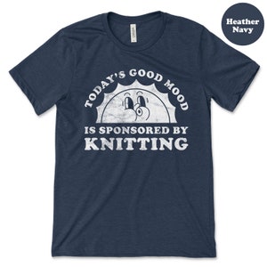 Op de afbeelding: Een heideblauwe t-shirt met een witte afbeelding van een lachende zon met de tekst "Today's Good Mood Is Sponsored By Knitting".