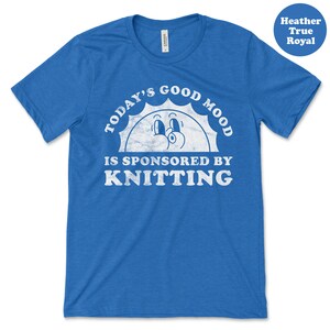 Op de afbeelding: Een koninklijk blauw t-shirt met een witte, verweerde afbeelding met de tekst "Today's Good Mood Is Sponsored By Knitting" met een cartoon zon gezicht.