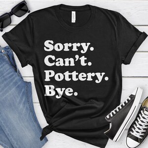 May include: Black t-shirt with the text "Sorry. Can't. Pottery. Bye." in white, sans-serif font. The shirt is styled with blue jeans, black sunglasses, and black and white sneakers. The shirt is knotted at the waist.