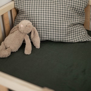 May include: A green crib sheet with a plush brown bunny toy and a gray and white gingham pillow. The crib is against a wall with a floral wallpaper featuring birds.