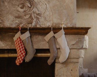 May include: Four Christmas stockings hang on a fireplace mantel. The stockings are made of fabric and have different patterns. One stocking is red and white plaid, one is gray and white plaid, and two are white with a subtle texture.