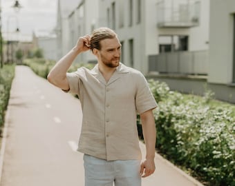 May include: A man wearing a light beige linen shirt with a collared neckline and short sleeves, light blue linen shorts, and black leather loafers. He is standing on a paved path with green bushes in the background.