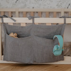May include: A black and white gingham fabric organiser with three pockets hangs on a wooden cot rail. The organiser has a wooden toy in one pocket and a pair of teal headphones in another pocket.