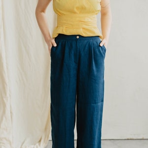 May include: A person wearing a yellow linen top and dark blue linen pants. The pants have a wide leg and a high waist. The person is standing on a white surface with lemons scattered around them.