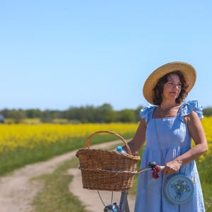 May include: A light blue sleeveless dress with ruffled shoulder straps, a straw hat, and a round light blue crossbody bag featuring a floral embroidery design. The outfit is paired with a bicycle equipped with a woven wicker basket on the handlebars.