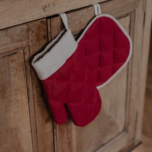 Peut inclure: Une manique de cuisine rouge matelassée avec une bordure blanche est accrochée à une porte d'armoire en bois.