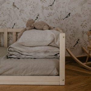 Peut inclure: Lit d'enfant avec un cadre en bois clair, avec une pile de literie aux tons neutres avec de fines rayures. Une peluche repose sur le dessus. Le papier peint présente un motif d'oiseaux et de branches.