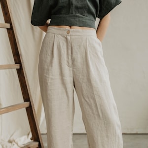 May include: Beige linen pants with a button closure and pleats. The pants are high-waisted and have a wide-leg design. The model is wearing a dark green top. A wooden ladder is in the background.