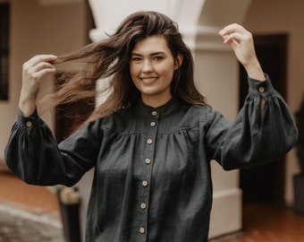 May include: A woman wearing a dark green, long-sleeved button-up shirt and matching skirt. The shirt has a high neckline and a gathered bodice. The skirt is a midi length and has a ruffled hem.