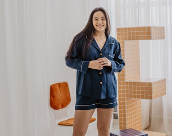 May include: A woman wearing a navy blue pajama set with white piping. The set includes a long-sleeved shirt and shorts. She is standing in a room with white walls and a window in the background.