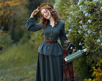 May include: A woman in a green dress and brown boots stands in a forest path. She is wearing a brown hat and holding a green thermos and a red and black plaid blanket.