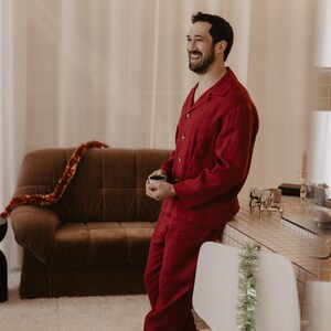 May include: A man in a red pyjama set smiles while holding a small cup. He is standing in a room with a brown sofa and a white chair with a green garland hanging from it.