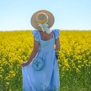 May include: A light blue sleeveless dress with ruffled shoulders, a straw hat with a cream-colored ribbon, and a round light blue crossbody bag featuring a floral embroidery design with a blue ribbon detail, worn in a field of yellow flowers.