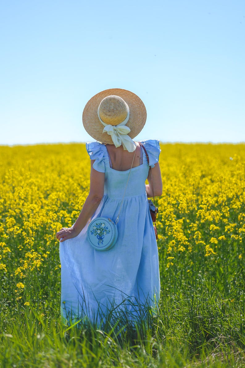 May include: A light blue sleeveless dress with ruffled shoulders, a straw hat with a cream-colored ribbon, and a round light blue crossbody bag featuring a floral embroidery design with a blue ribbon detail, worn in a field of yellow flowers.