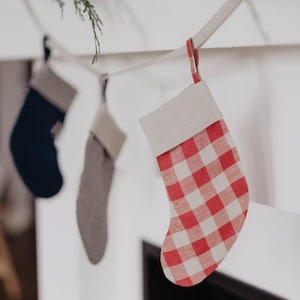 Op de afbeelding: Drie kerstsokken hangen aan een witte schoorsteenmantel. De dichtstbijzijnde sok heeft een rood-wit geblokt patroon. De andere twee sokken zijn grijs en marineblauw. Boven de sokken is groen te zien.