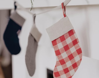 May include: Three Christmas stockings hang from a white mantel. The closest stocking has a red and white checkered pattern. The other two stockings are gray and navy blue. Greenery is visible above the stockings.