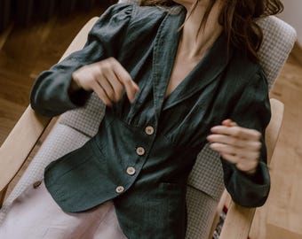 May include: A woman wearing a dark green linen blazer with buttons and a light pink linen pants. She is sitting in a chair with a white and gray patterned fabric.