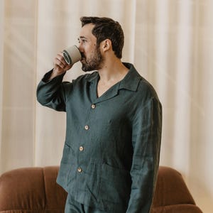 May include: A person wearing a dark green linen pyjama set with a button-down shirt and matching trousers. The person is standing in front of a brown sofa and drinking from a white ceramic mug.