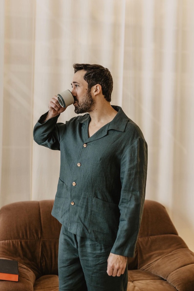 May include: A person wearing a dark green linen pyjama set with a button-down shirt and matching trousers. The person is standing in front of a brown sofa and drinking from a white ceramic mug.