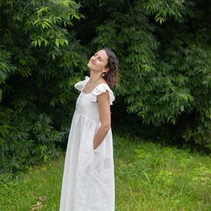 May include: A floor-length white dress featuring a square neckline, ruffled cap sleeves, and a gathered waistline. The garment has a loose, flowing silhouette and includes side pockets, crafted from a solid, opaque white fabric.