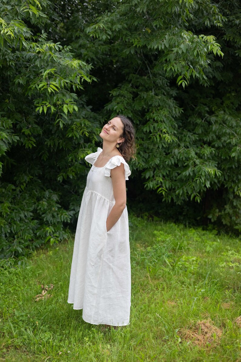 May include: A floor-length white dress featuring a square neckline, ruffled cap sleeves, and a gathered waistline. The garment has a loose, flowing silhouette and includes side pockets, crafted from a solid, opaque white fabric.