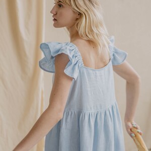 May include: A light blue sleeveless dress featuring a square neckline and ruffled cap sleeves. The garment has a gathered waistline that creates a loose, flowing silhouette. The fabric appears to have a textured, woven finish throughout the design.