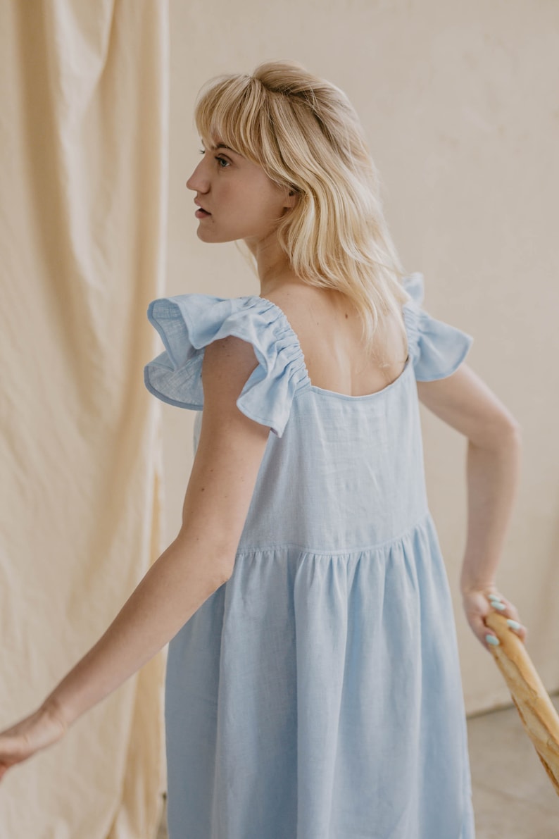 May include: A light blue sleeveless dress featuring a square neckline and ruffled cap sleeves. The garment has a gathered waistline that creates a loose, flowing silhouette. The fabric appears to have a textured, woven finish throughout the design.